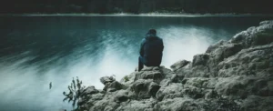 a man sits on a rock staring across a lake at dusk. 7 síntomas de la dependencia emocional